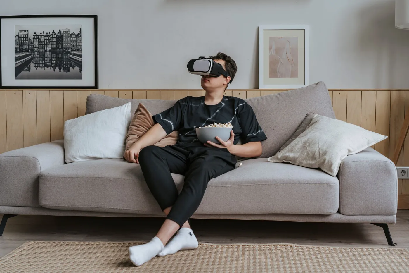 Man sitting on a gray sofa in a living room wearing a VR headset and holding a bowl of popcorn