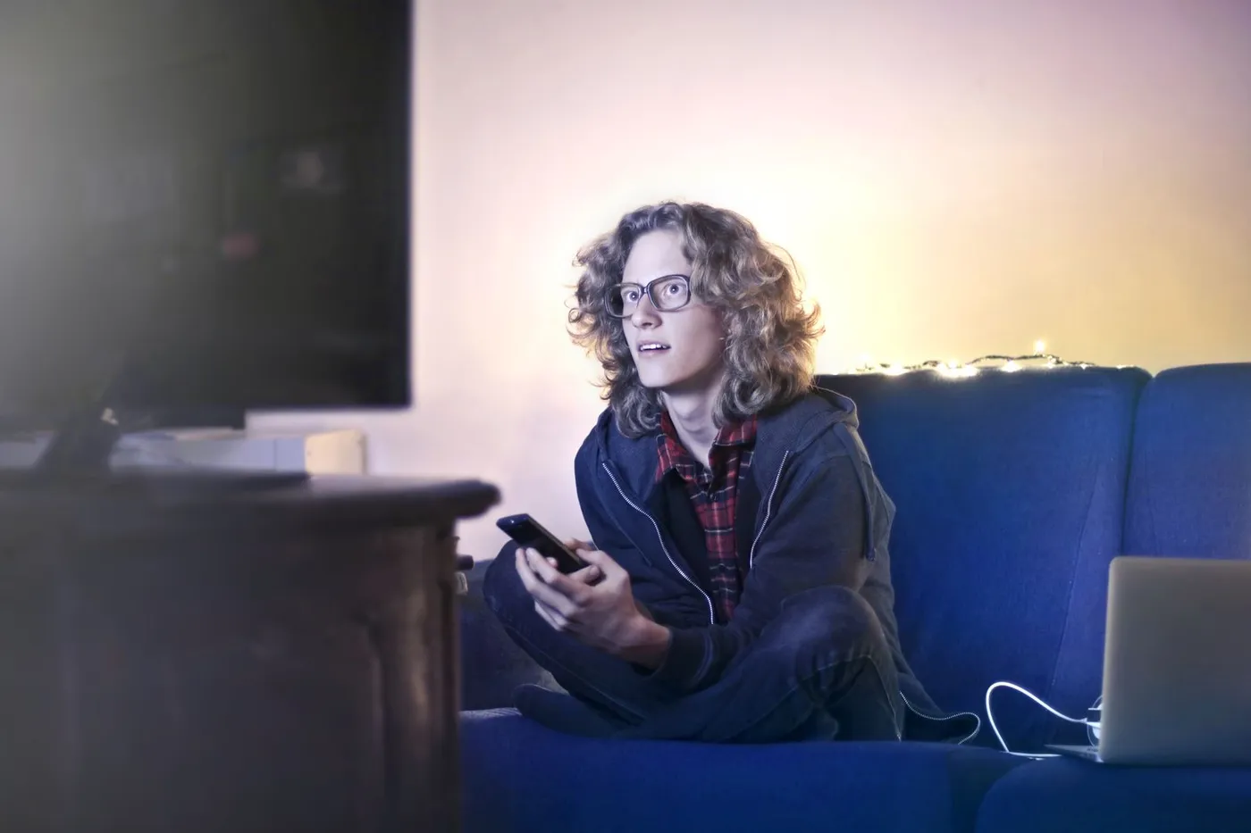 Young man sitting cross-legged on a blue sofa holding a smartphone, with a TV in the foreground and an open laptop beside him
